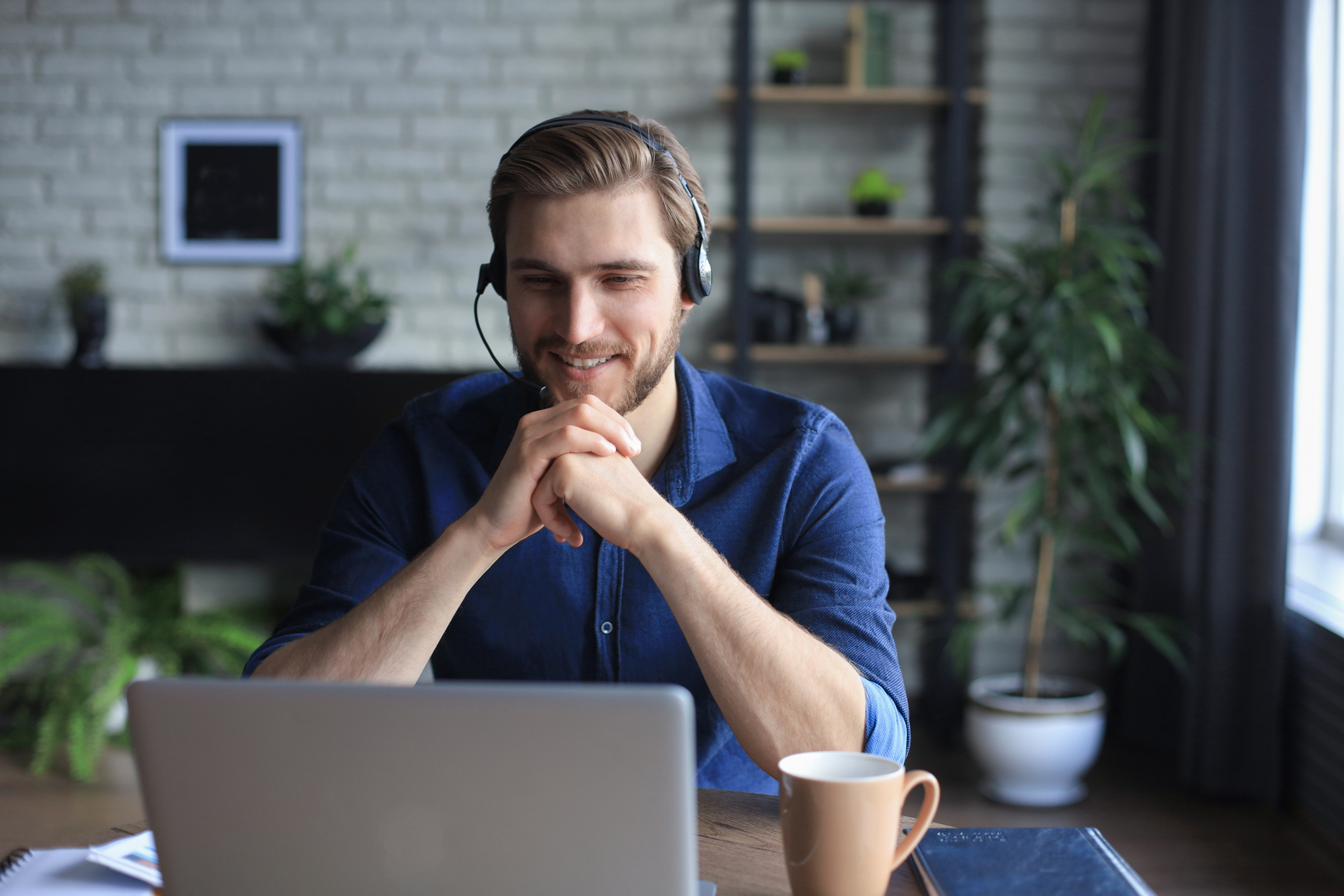 Man met hoofdtelefoon zit glimlachend achter zijn laptop in een huiselijke kantooromgeving met planten en een kop koffie op tafel.