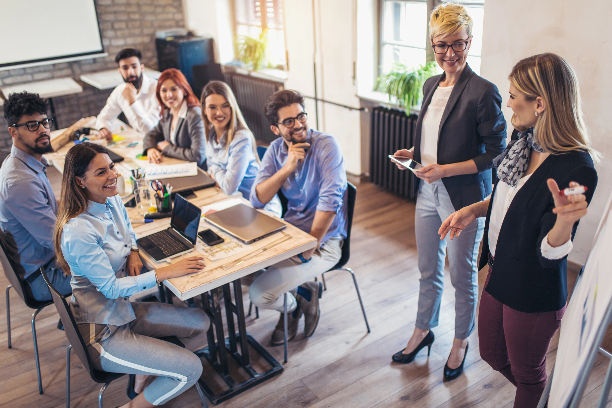 Een diverse groep professionals in een kantooromgeving tijdens een vergadering. Zeven mensen zitten aan een tafel met laptops en notitieboeken, terwijl twee vrouwen staan en de discussie leiden. De sfeer oogt positief en betrokken, met mensen die glimlachen en aandachtig luisteren.