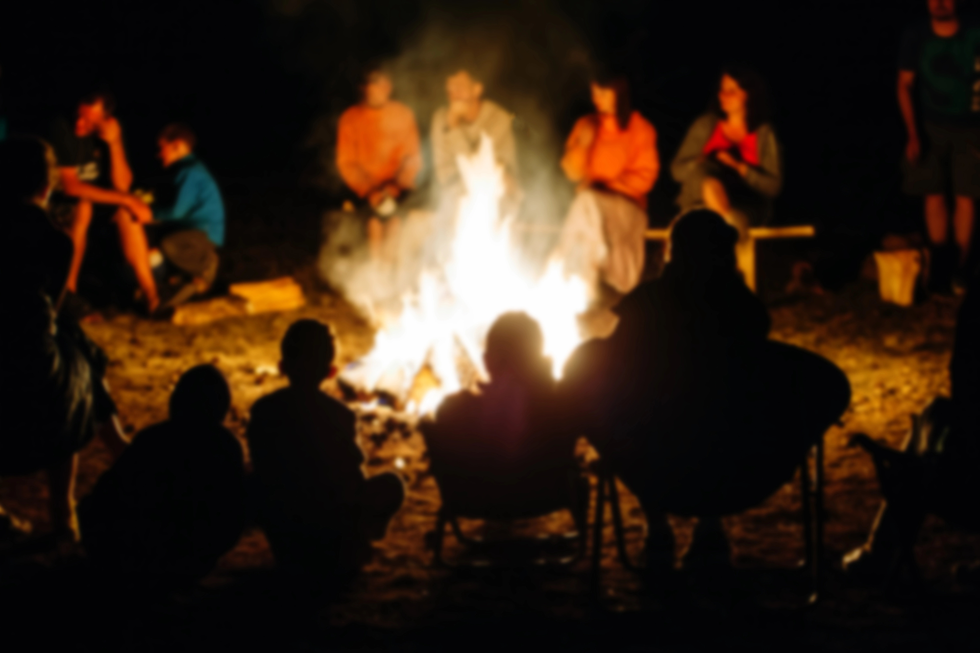 Een groep mensen zit 's nachts in een cirkel rond een groot kampvuur. De silhouetten van enkele personen zijn zichtbaar op de voorgrond, terwijl anderen in felle kleuren gekleed zijn aan de overkant van het vuur. Het vuur verlicht de omgeving met een warme gloed.