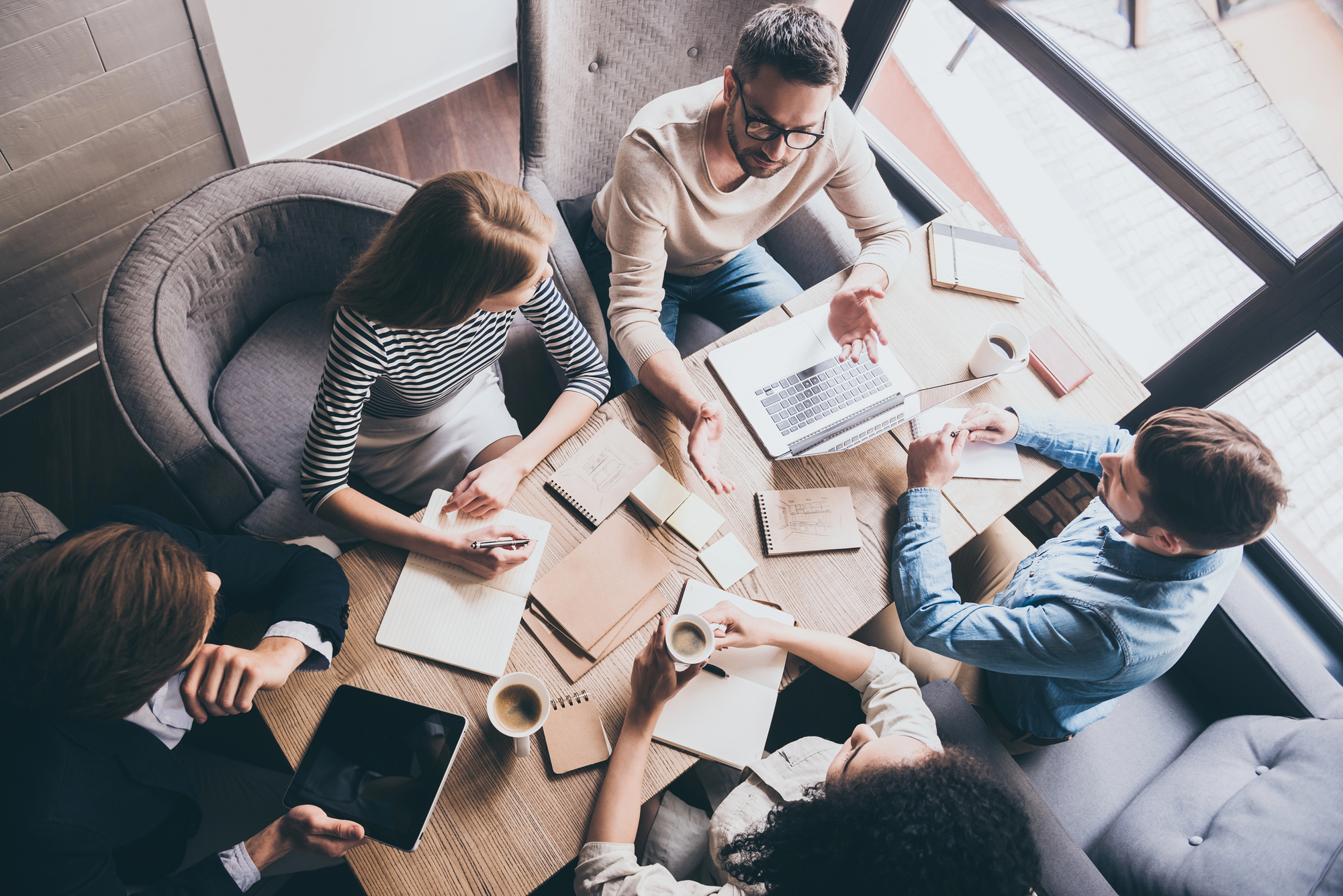 Een bovenaanzicht van een informele vergadering waarbij vijf mensen rond een houten tafel zitten. Op tafel liggen diverse notitieboeken, papieren en een laptop. Er staan ook enkele koppen koffie. Twee personen dragen casual kleding - één in een beige trui met bril, een ander in een gestreept shirt. De sfeer oogt ontspannen en collaboratief, met mensen die actief lijken te discussiëren en aantekeningen maken. De setting lijkt een modern kantoor of werkruimte met natuurlijk licht dat binnenvalt door een raam.