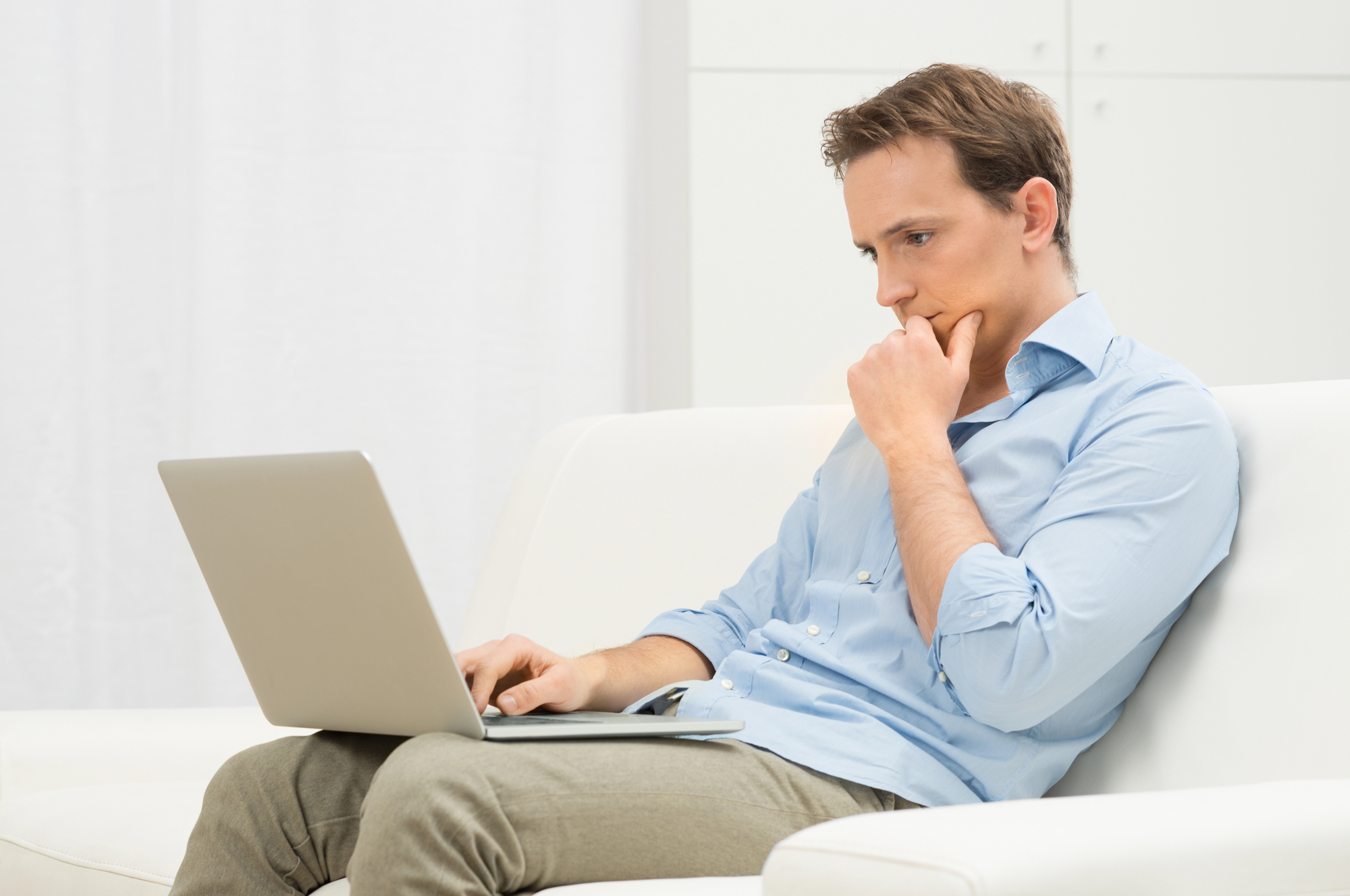 Man in lichtblauw overhemd zit op een witte bank met een laptop op schoot, hand bij zijn kin, kijkend met een peinzende blik terwijl hij een beslissing overweegt.