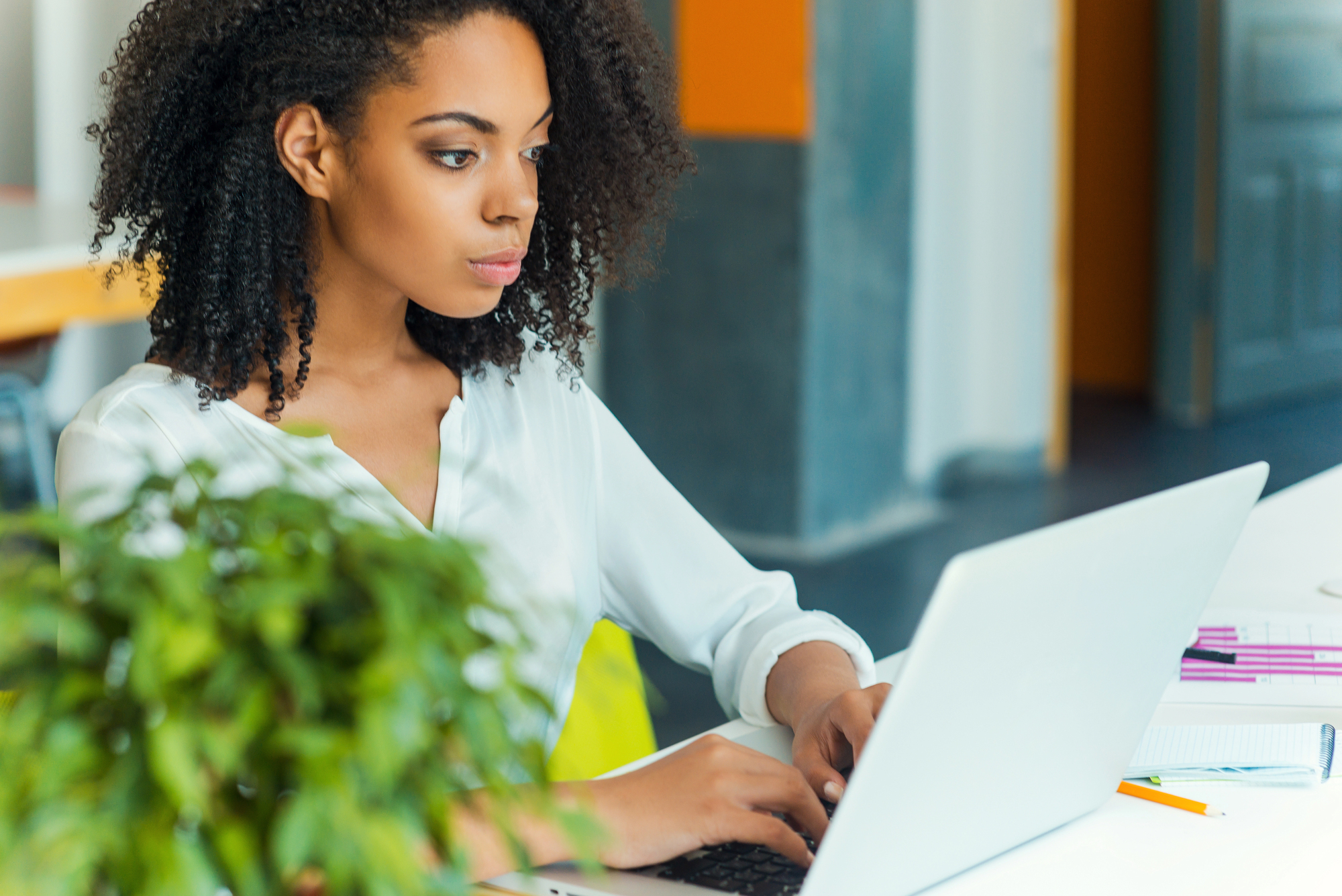 Een jonge vrouw met krullend haar zit geconcentreerd te werken op een laptop. Ze draagt een witte blouse en zit in een moderne kantooromgeving. Op de voorgrond is een groene plant vaag zichtbaar.