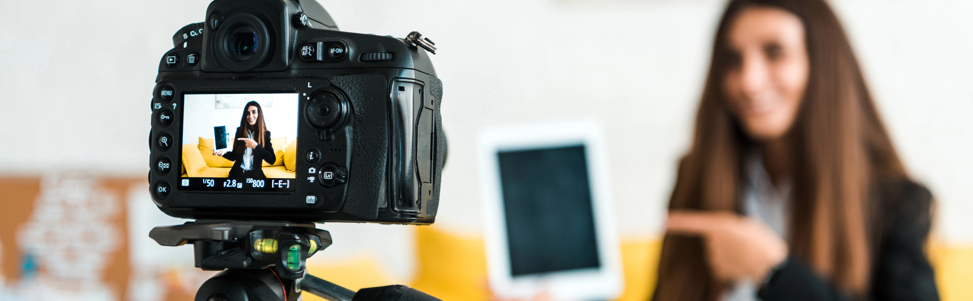 “Een vrouw met lang bruin haar presenteert een tablet terwijl ze voor een camera staat. De camera is scherpgesteld op het scherm, waarop de vrouw zichtbaar is terwijl ze naar de tablet wijst. De achtergrond is onscherp en bevat gele elementen.”