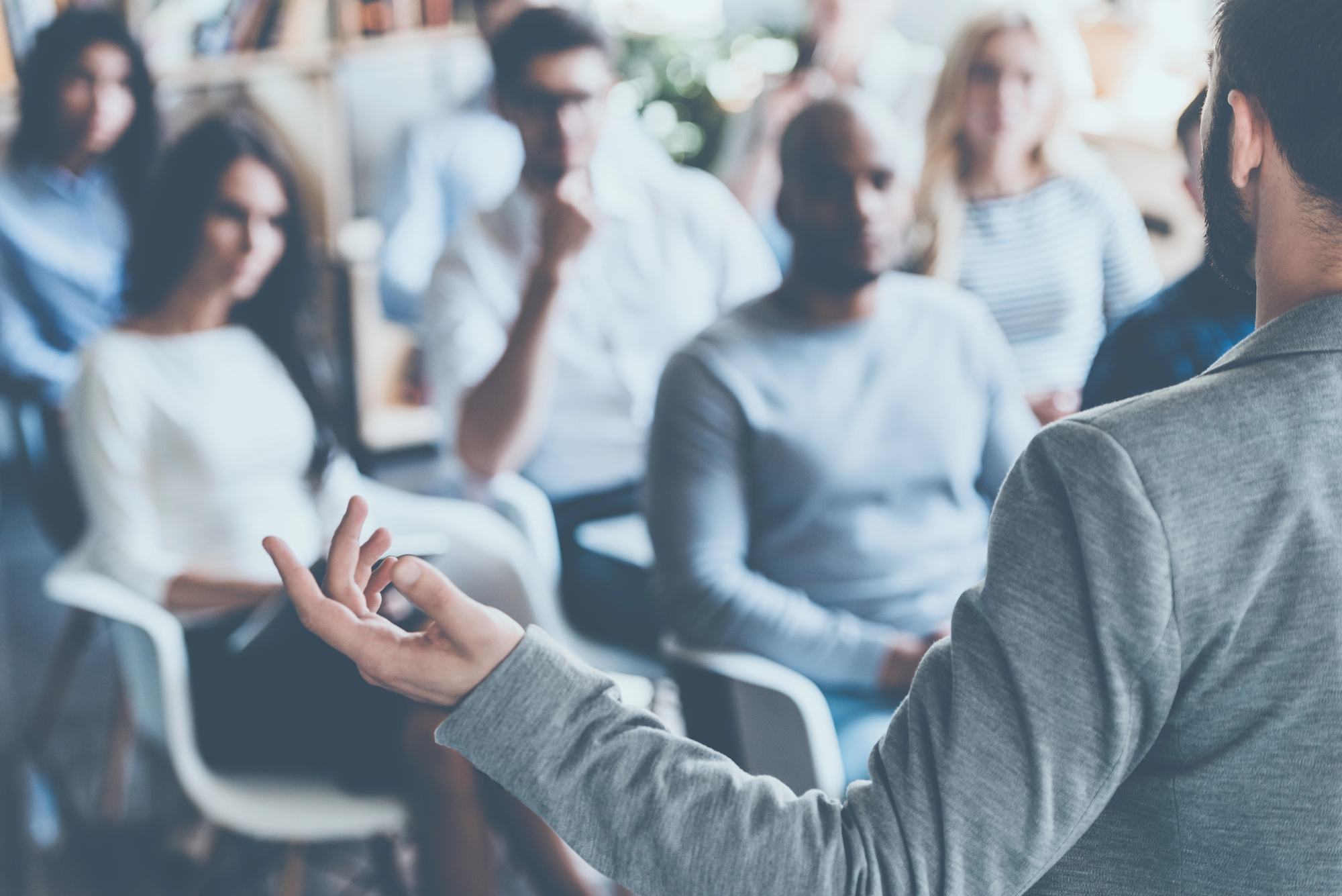 Een groep mensen luistert aandachtig naar een spreker tijdens een presentatie of workshop. De spreker, gezien vanaf de rug, gebaart met zijn hand terwijl hij praat. De toehoorders, zittend in moderne stoelen, kijken gefocust naar de spreker.