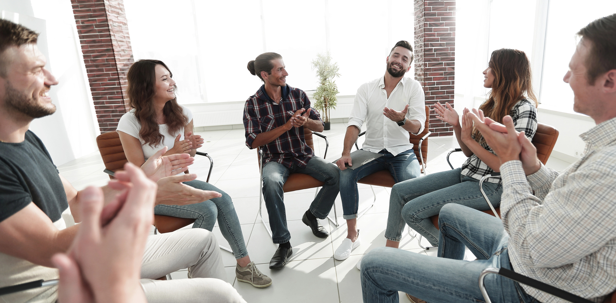 Een groep mensen zit in een kring. Ze lachen en applaudisseren. De ruimte is licht en modern, met grote ramen en bakstenen muren. Iedereen zit op stoelen in een open opstelling.