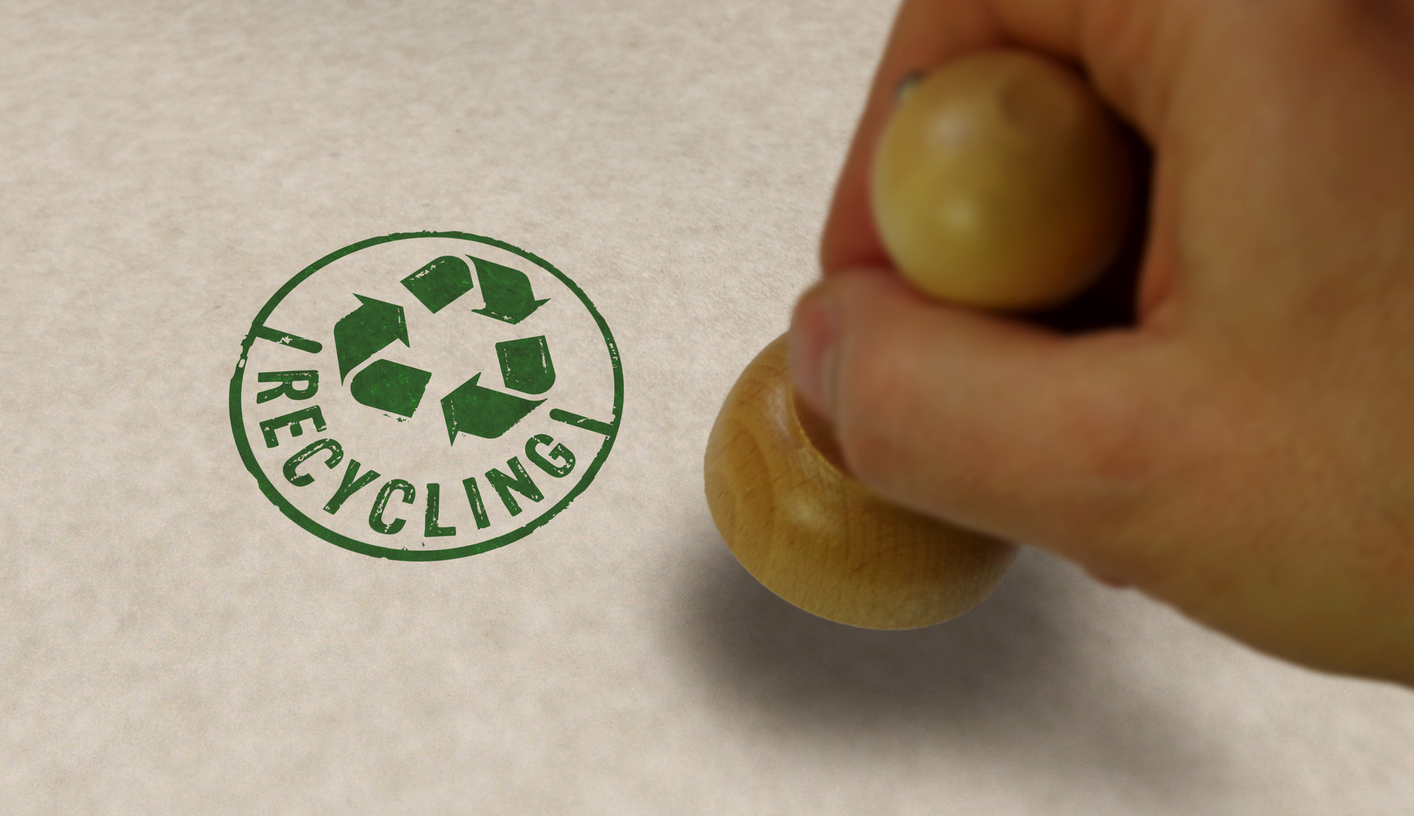 Een foto van een hand die een stempel op papier drukt. De stempel bevat een groen recycling symbool en het woord “RECYCLING” eronder.