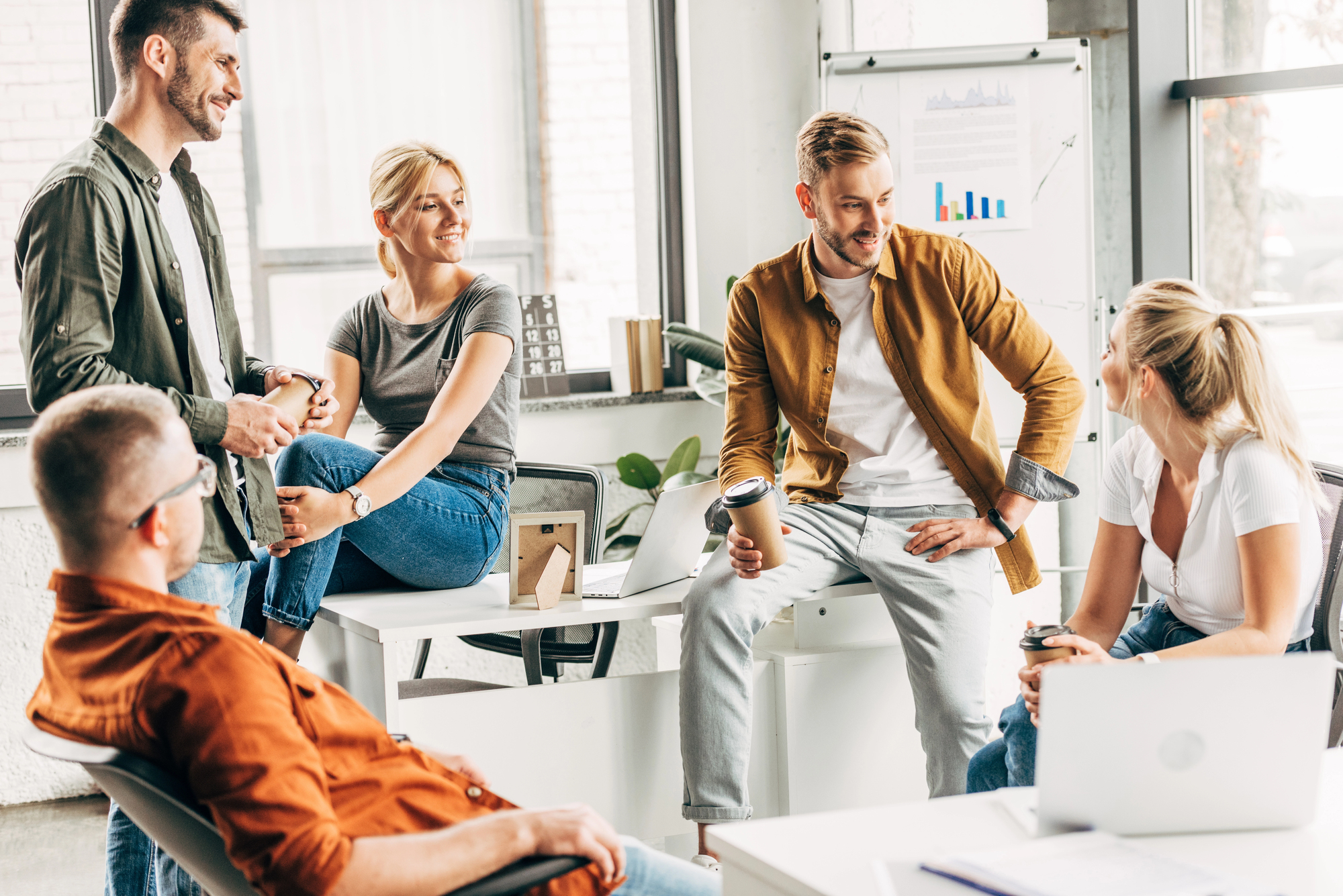 Groep jonge professionals in casual kleding die informeel samenkomt in een lichte kantoorruimte, met een whiteboard op de achtergrond dat grafieken toont. Ze lijken betrokken in een ontspannen discussie of brainstormsessie.