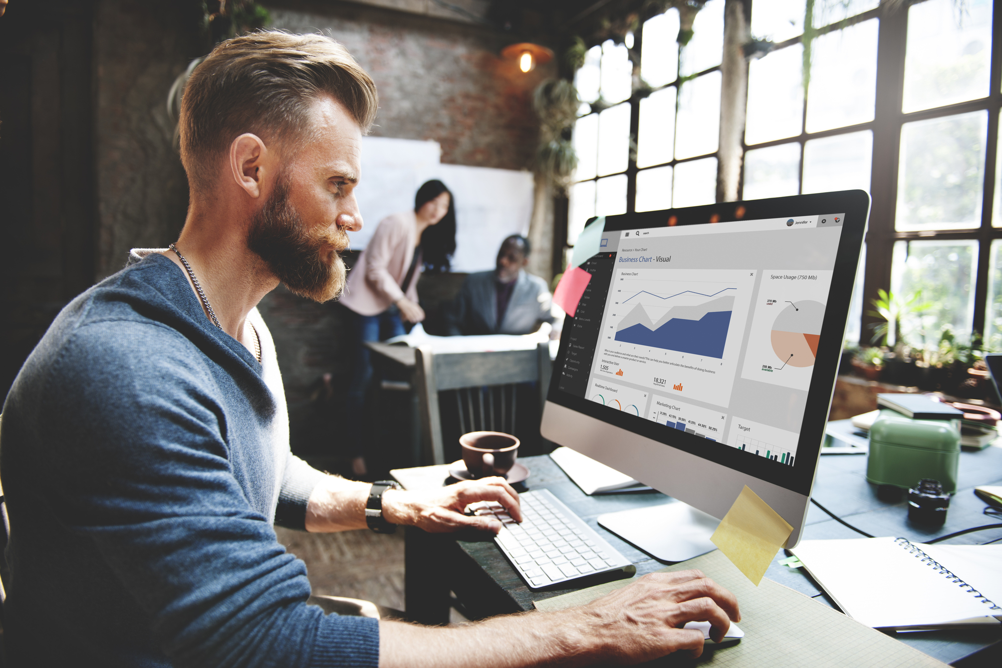 Een man met een baard en kort, gestyled haar die gefocust werkt op een computer in een moderne kantooromgeving. Hij draagt een blauwe trui en typt op een toetsenbord terwijl hij naar een groot computerscherm kijkt met daarop verschillende grafieken en data-analyses. Op de achtergrond zijn een man en een vrouw te zien die samen overleggen bij een tafel met papieren. Het kantoor heeft grote ramen die veel licht binnenlaten en de ruimte heeft een industriële uitstraling met bakstenen muren en planten.