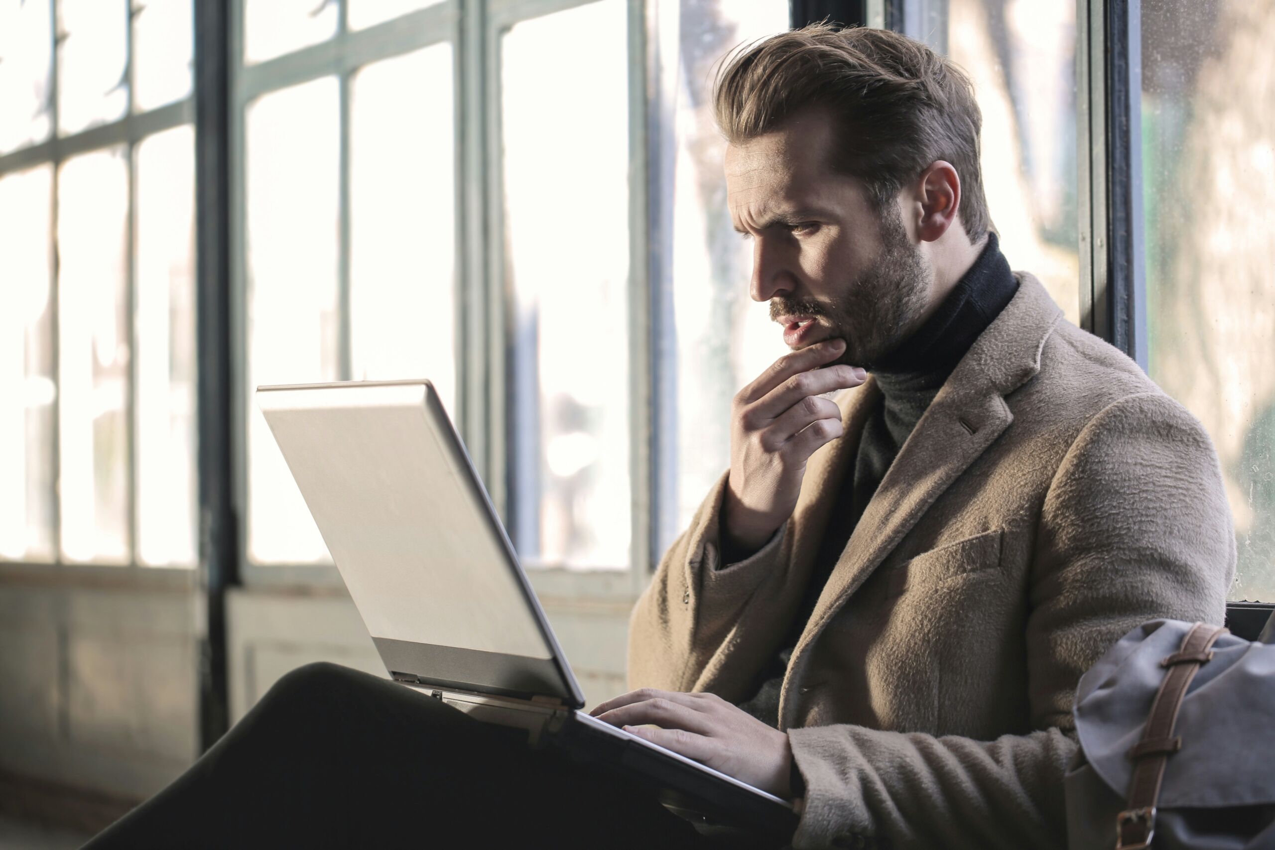 Een man in een beige jas zit bij een raam en kijkt met een peinzende uitdrukking naar zijn laptop. Hij heeft zijn hand tegen zijn kin, wat suggereert dat hij diep in gedachten is of een probleem probeert op te lossen. Het natuurlijke licht van het raam valt op hem, wat een contemplatieve sfeer creëert. Zijn kleding en houding geven een professionele indruk.