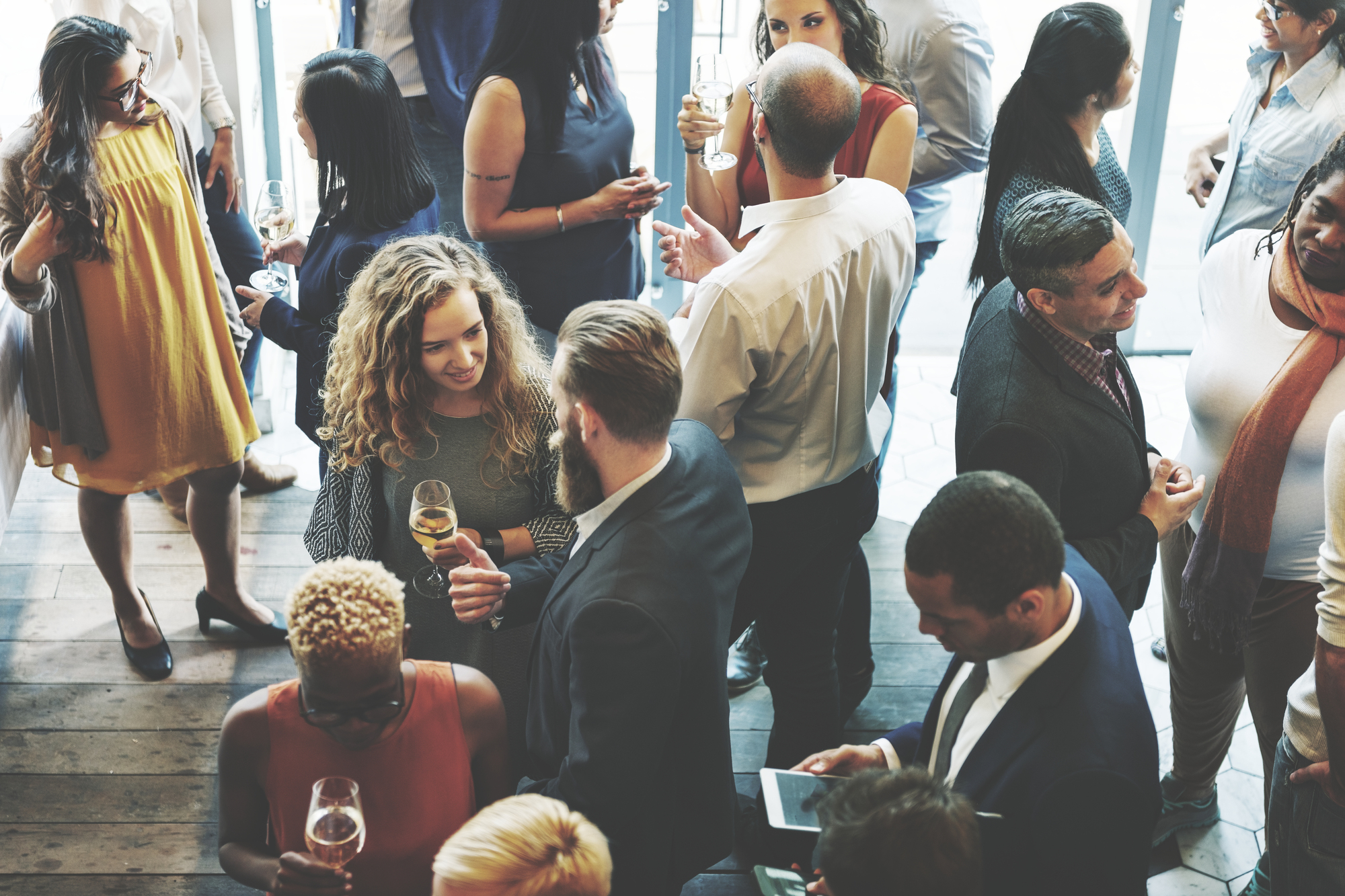 Diverse groep professionals in zakelijke kleding socialiseert tijdens een netwerkborrel, met wijnglazen in de hand in een ruimte met grote ramen en houten vloer.