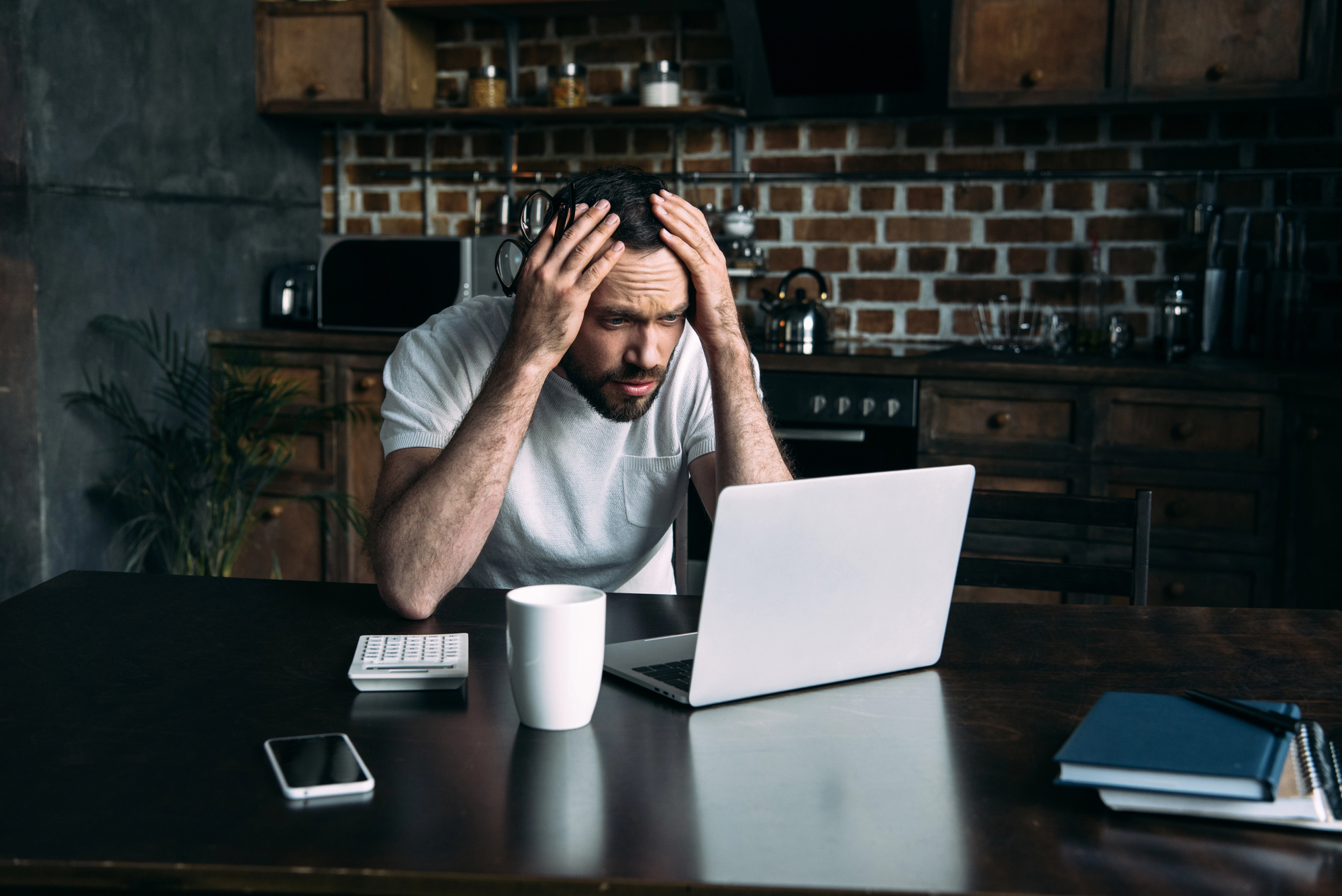 Een man in een witte t-shirt zit aan een tafel in een keuken, met zijn handen op zijn hoofd terwijl hij met een gefrustreerde blik naar een laptop kijkt. Op tafel staan ook een koffiekop, rekenmachine, smartphone en notitieboek.