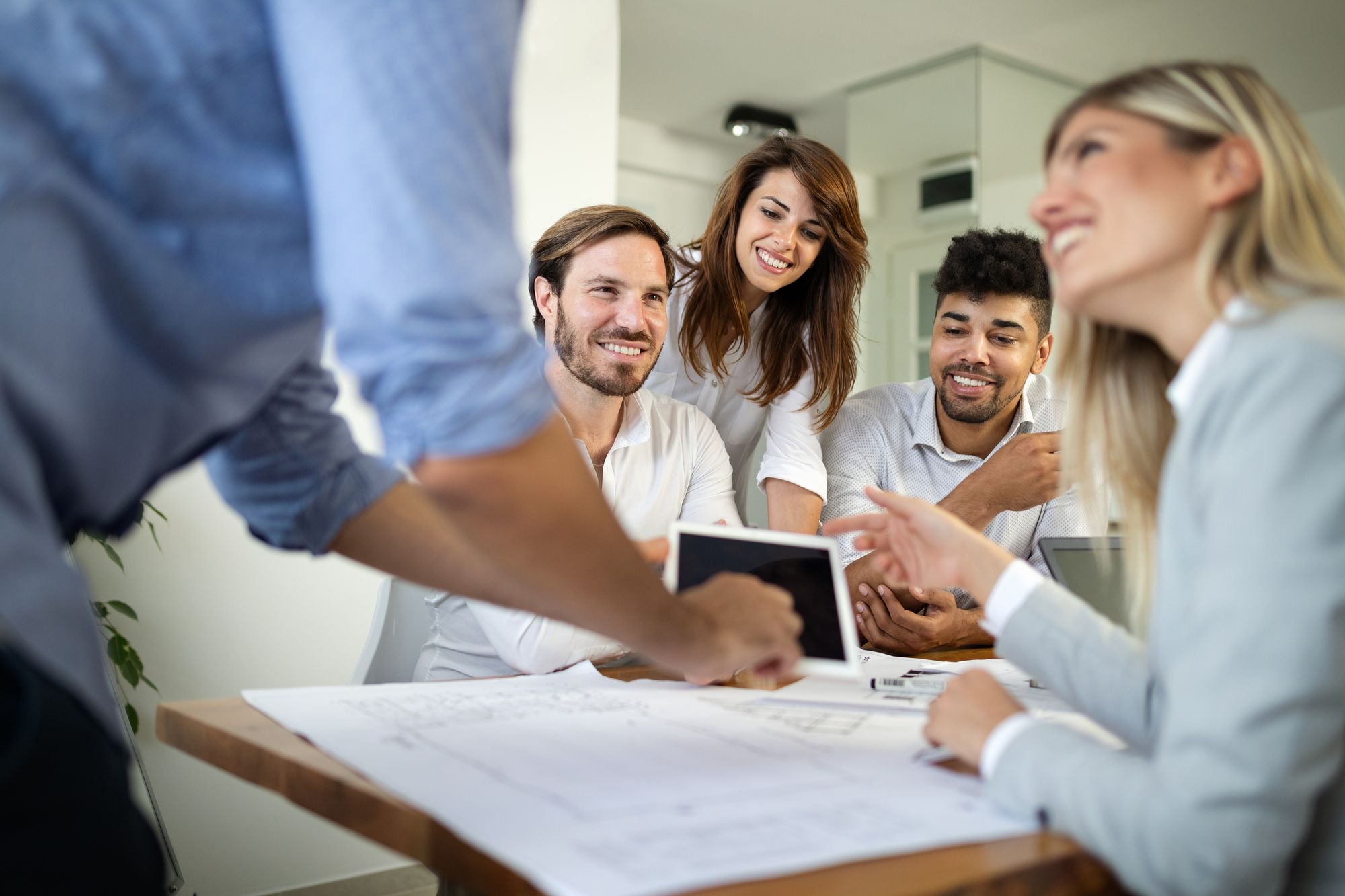 Een diverse groep lachende jonge professionals in een revenue team meeting bekijkt samen documenten en tablets aan een vergadertafel.