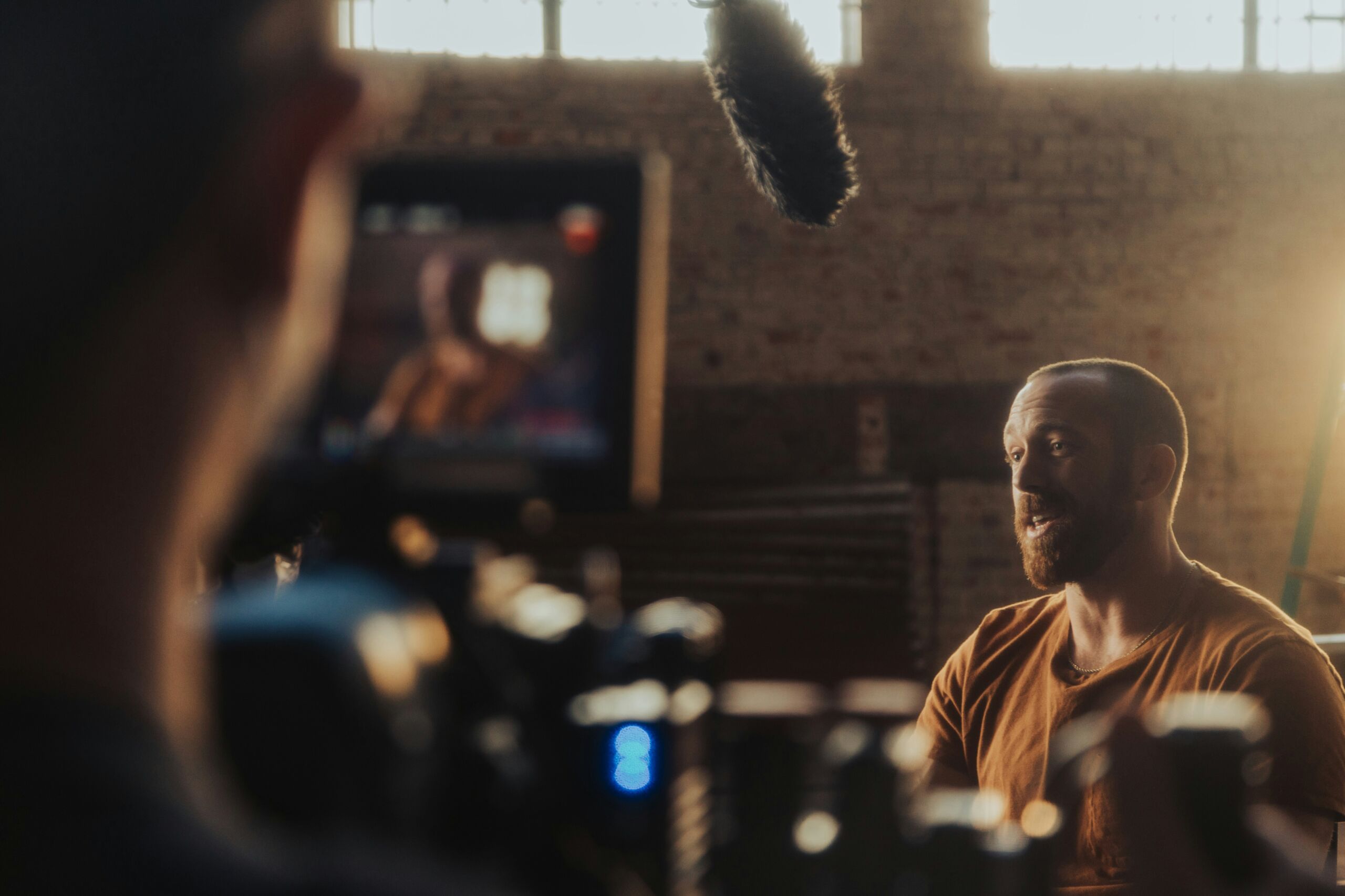 Man met baard in oranje shirt wordt geïnterviewd voor camera in studio met natuurlijk licht en zichtbare microfoonboom