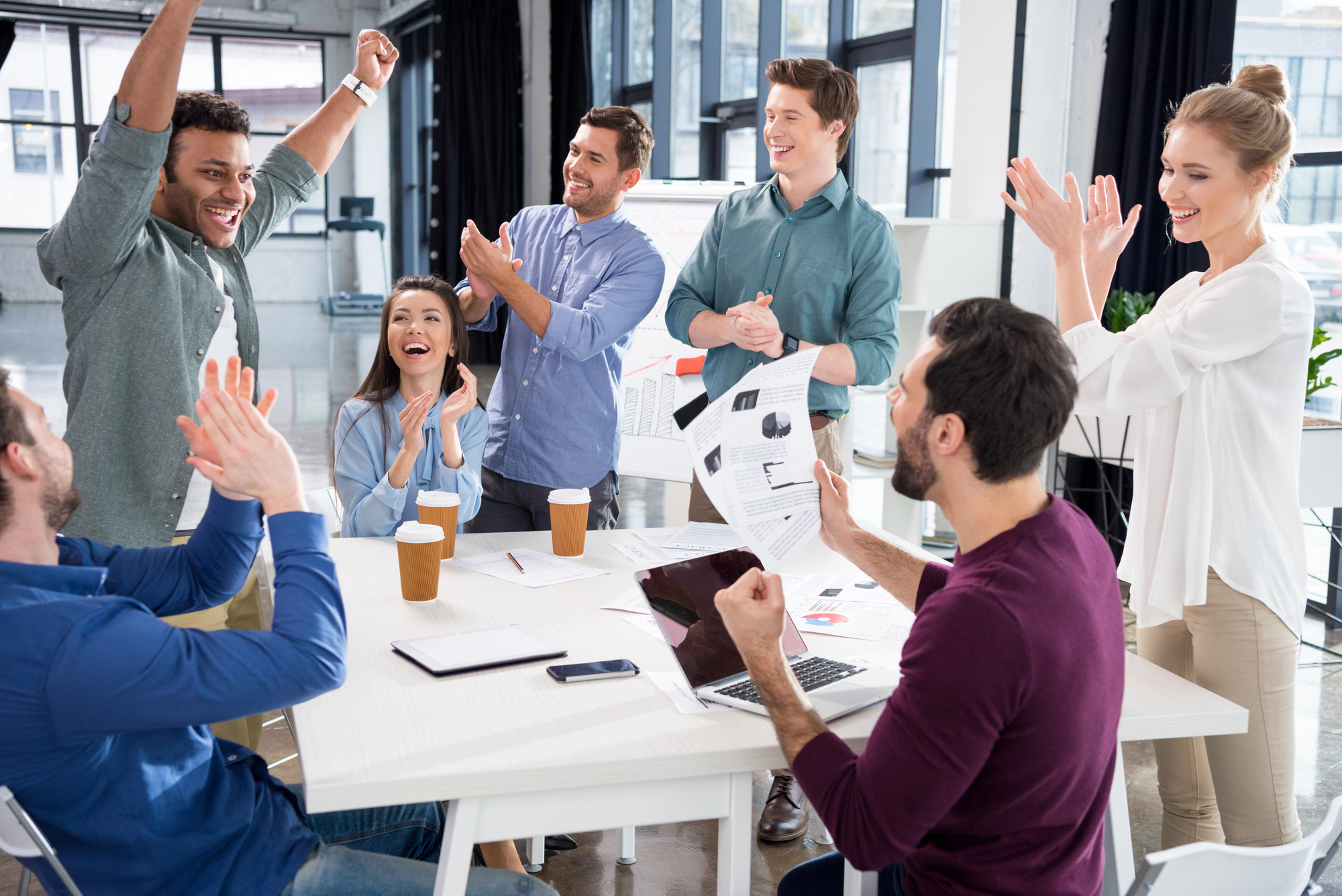 Team viert succes tijdens vergadering, diverse medewerkers juichen en klappen rondom een vergadertafel met laptops en documenten in een modern kantoor