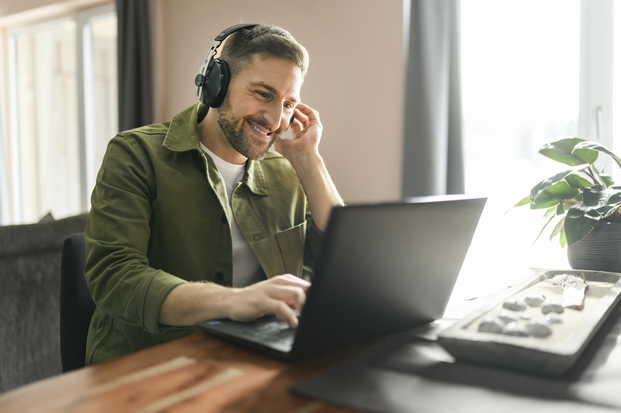 Man in groen overhemd zit achter laptop en glimlacht. Hij draagt een headset met microfoon en zit in een lichte ruimte.