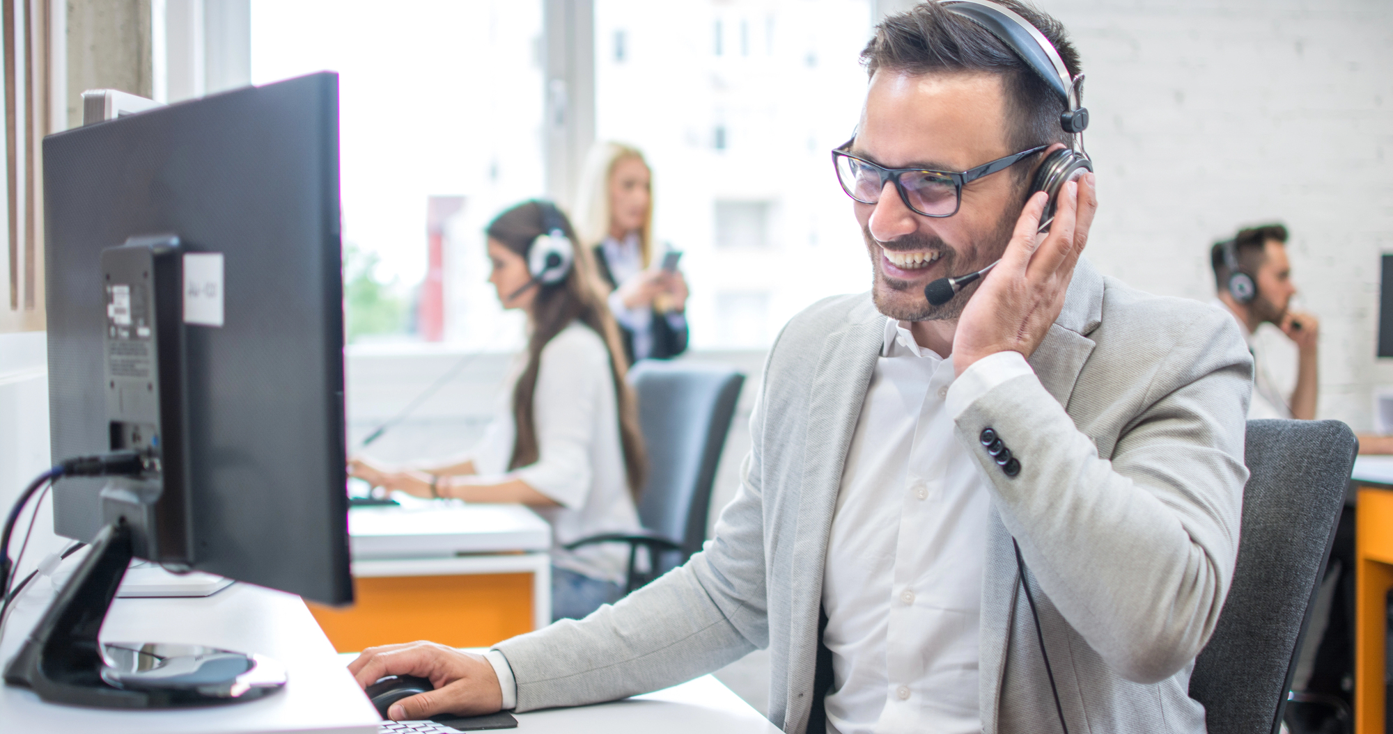 Glimlachende salesprofessional in grijs colbert en headset achter computerscherm in open kantoorruimte, met collega's op de achtergrond.