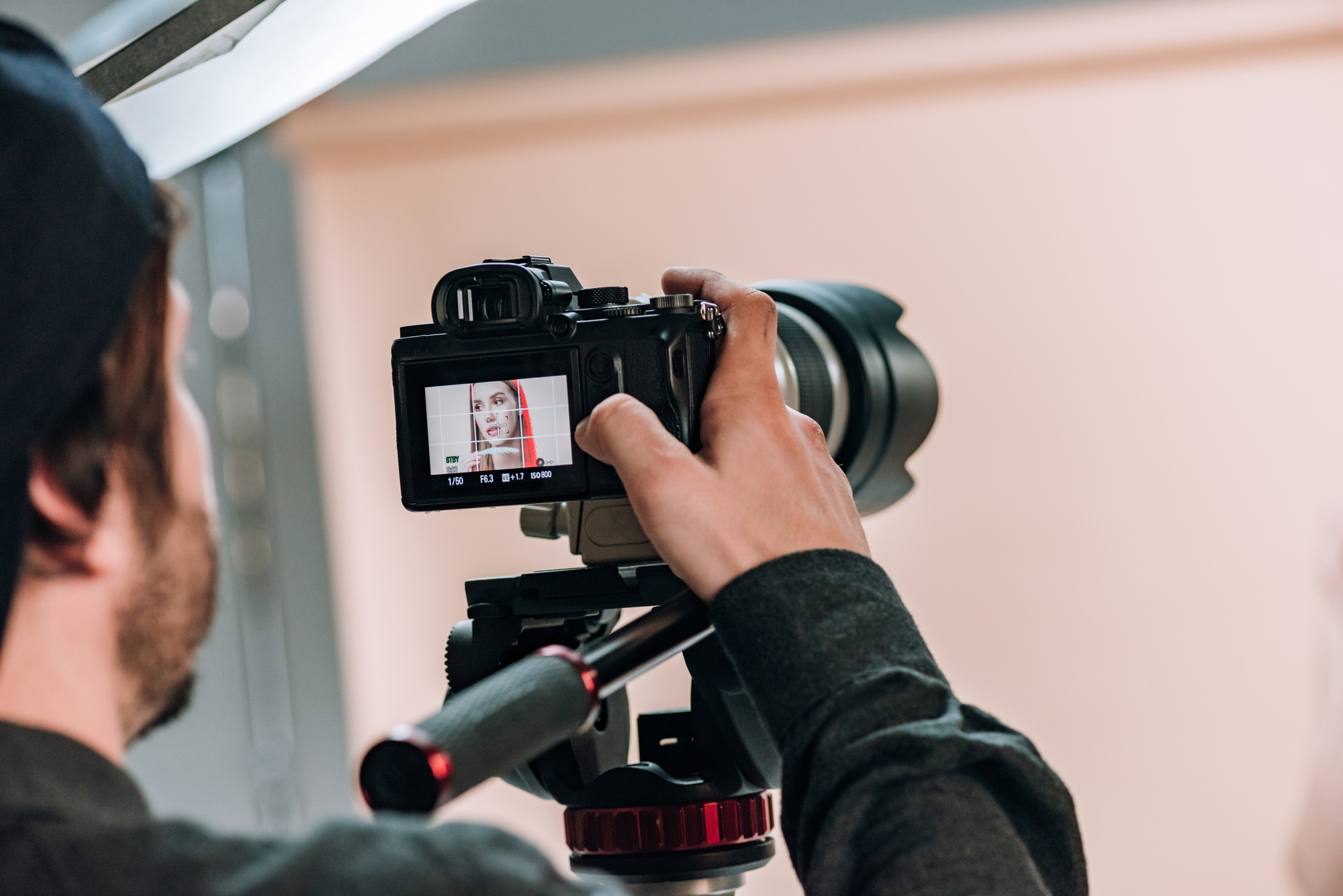 Close-up van een cameraman die een video-opname maakt. Op het display van de camera is een vrouw te zien met een rode sjaal, scherp in beeld gebracht. De cameraman draagt een donker kledingstuk en een pet, terwijl hij de camera vasthoudt en de focus aanpast. De achtergrond is neutraal en licht van kleur.