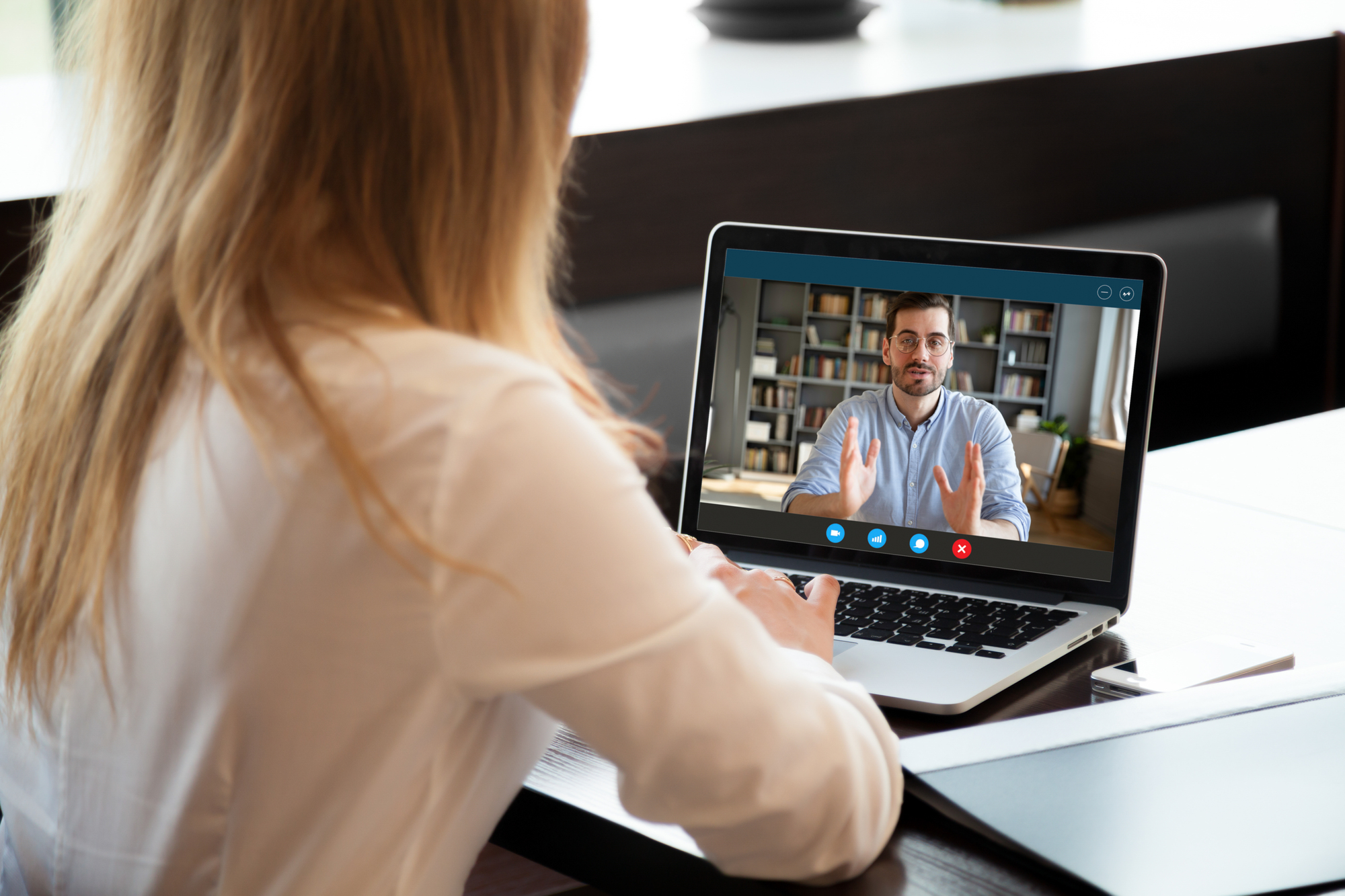 Een vrouw in een wit shirt kijkt naar een videogesprek op haar laptop, waarin een man met bril en baard in een lichte trui voor een boekenkast zit te praten en handgebaren maakt.