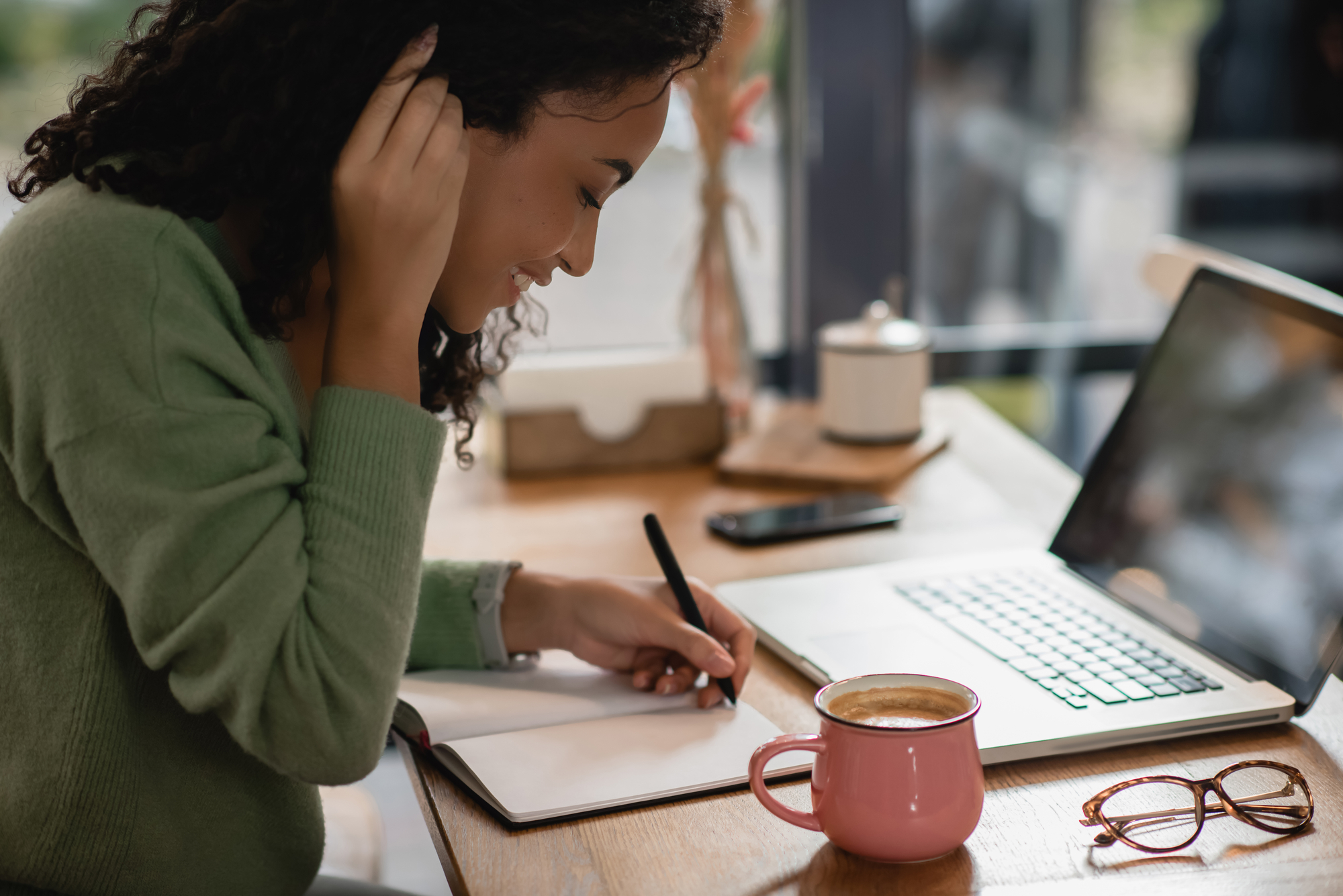 Een jonge vrouw met krullend haar zit aan een houten tafel in een café. Ze draagt een groene trui en schrijft in een notitieboekje met een pen. Voor haar staan een laptop en een roze koffiekop. Haar bril en smartphone liggen ook op de tafel.